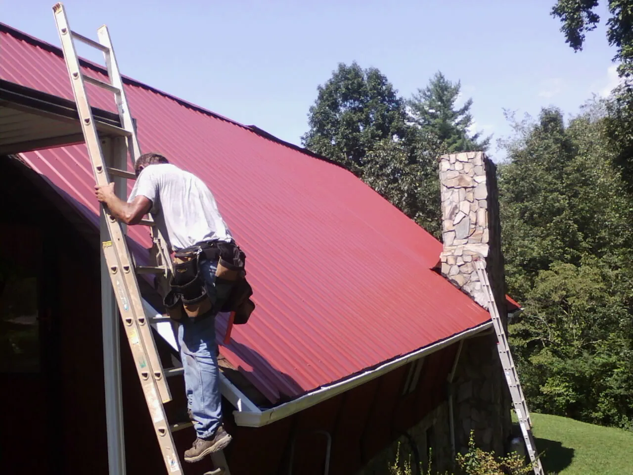 Roofer on a ladder beside a red metal roof and stone chimney — Soffit & Fascia Repair by Roofing Brothers in South Milwaukee